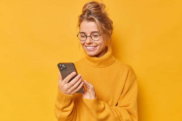 modern woman with combed hair holds mobile phone reads screen chats glad receive pleasant message wears round spectacles casual jumper isolated yellow background downloads news application